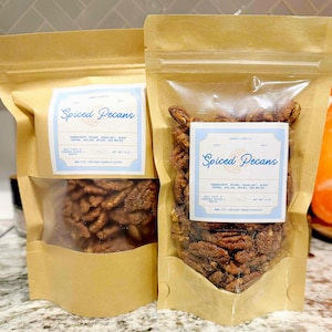 Puede incluir: Dos bolsas de papel kraft llenas de nueces pecanas especiadas. Cada bolsa tiene una ventana transparente que muestra las nueces. Las bolsas tienen una etiqueta azul claro con las palabras "Spiced Pecans". Los ingredientes están listados.