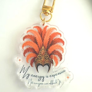 Acrylic keychain with showgirl hat design and “My Energy Is Expensive” quote – T Swift Life of a Showgirl inspired statement accessory. Close up of acrylic keychain featuring glamorous showgirl headdress and fancy “My Energy Is Expensive” lettering – glossy statement accessory for keys or bags. Showgirl headdress design, pop culture inspired accessory for Swiftie fans.