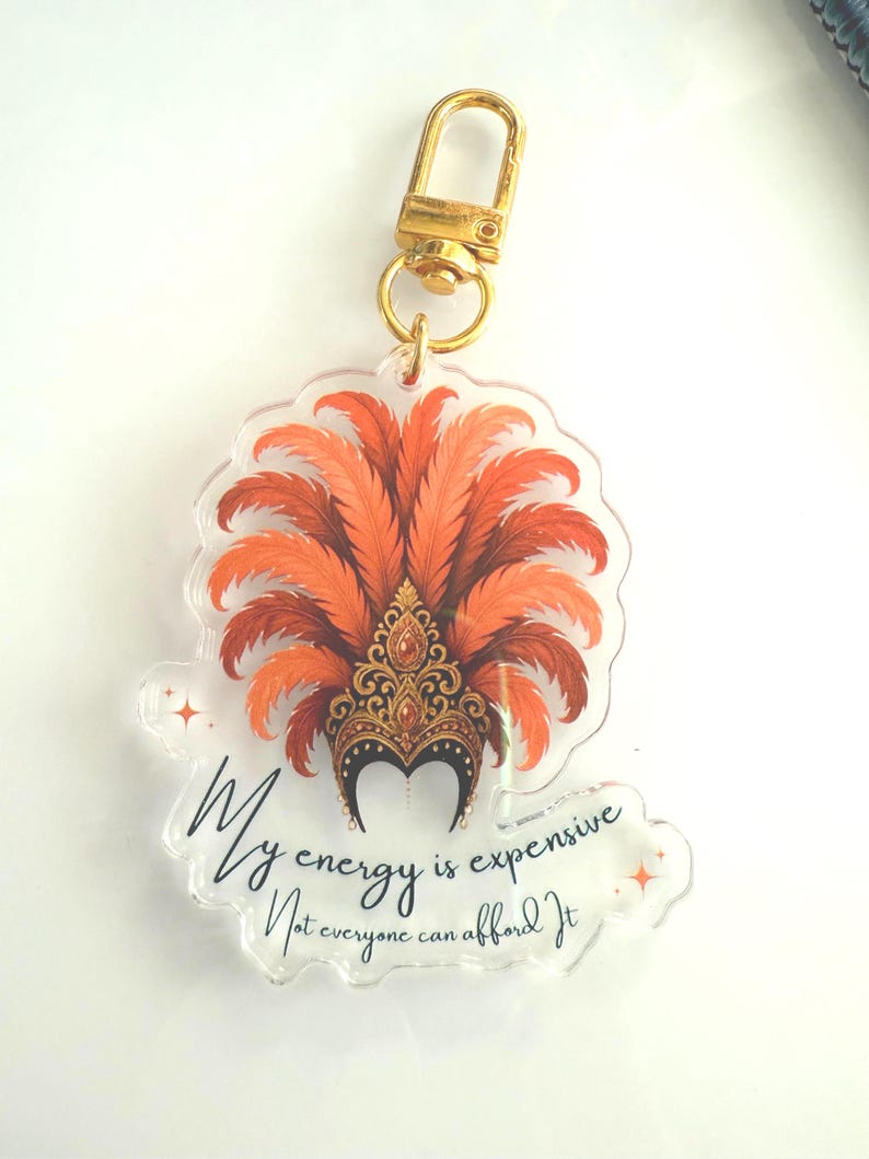 Acrylic keychain with showgirl hat design and “My Energy Is Expensive” quote – T Swift Life of a Showgirl inspired statement accessory. Close up of acrylic keychain featuring glamorous showgirl headdress and fancy “My Energy Is Expensive” lettering – glossy statement accessory for keys or bags. Showgirl headdress design, pop culture inspired accessory for Swiftie fans.