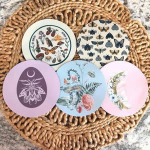 May include: Five round coasters with nature-themed designs. One coaster features an owl and butterflies, another has a moth pattern, and others showcase mushrooms, flowers, and a crescent moon. The coasters are arranged on a woven mat.