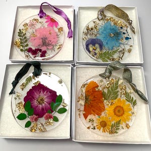 Könnte beinhalten: Vier klare, runde Ornamente mit getrockneten Blumen und Goldblatt-Akzenten, jeweils in einer weißen Box. Die Ornamente zeigen rosa, blaue, lila und gelbe Blumen mit passenden Bändern. Eine einzigartige Blumendekoration.