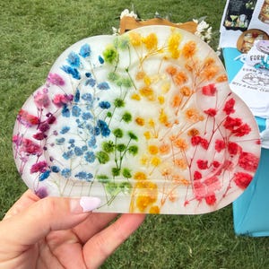 May include: A cloud-shaped resin tray featuring a rainbow gradient of embedded dried baby's breath flowers. The translucent material displays a spectrum of pink, blue, green, yellow, orange, and red floral stems arranged in a fan-like pattern.