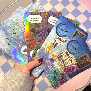 May include: A hand holding a collection of holographic postcards. The postcards feature various designs, including a moose, maple leaf, hockey stick, and a crescent moon. Text includes "I'm coming to the cottage" and "Brooklyn NYC".