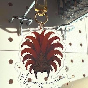 Acrylic keychain with showgirl hat design and “My Energy Is Expensive” quote – T Swift Life of a Showgirl inspired statement accessory. Close up of acrylic keychain featuring glamorous showgirl headdress and fancy “My Energy Is Expensive” lettering – glossy statement accessory for keys or bags. Showgirl headdress design, pop culture inspired accessory for Swiftie fans.