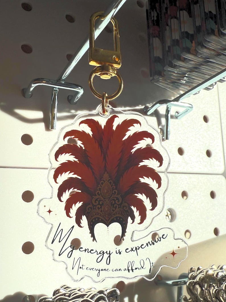 Acrylic keychain with showgirl hat design and “My Energy Is Expensive” quote – T Swift Life of a Showgirl inspired statement accessory. Close up of acrylic keychain featuring glamorous showgirl headdress and fancy “My Energy Is Expensive” lettering – glossy statement accessory for keys or bags. Showgirl headdress design, pop culture inspired accessory for Swiftie fans.