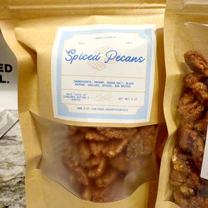 May include: A brown paper bag with a clear window filled with spiced pecans. The label reads "Spiced Pecans" with ingredients listed: pecans, sugar, salt, black pepper, chilies, spices, and egg whites. Net weight is 8 oz.