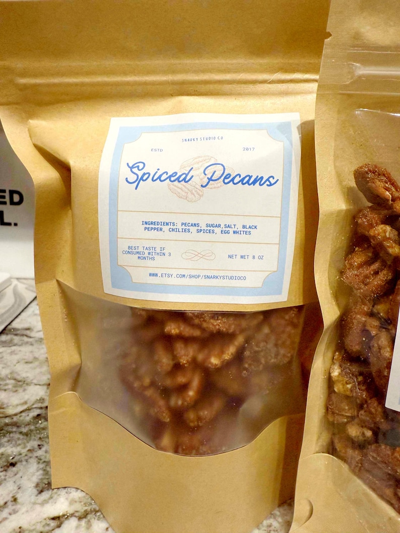 May include: A brown paper bag with a clear window filled with spiced pecans. The label reads "Spiced Pecans" with ingredients listed: pecans, sugar, salt, black pepper, chilies, spices, and egg whites. Net weight is 8 oz.
