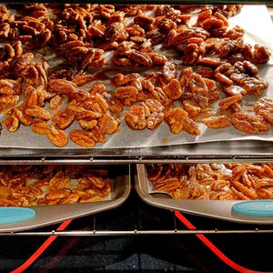 May include: Oven-baked pecans on parchment paper, with more pecans on trays below. The nuts are golden brown and appear glazed, suggesting a sweet coating. The oven's interior is visible, with red heating elements.
