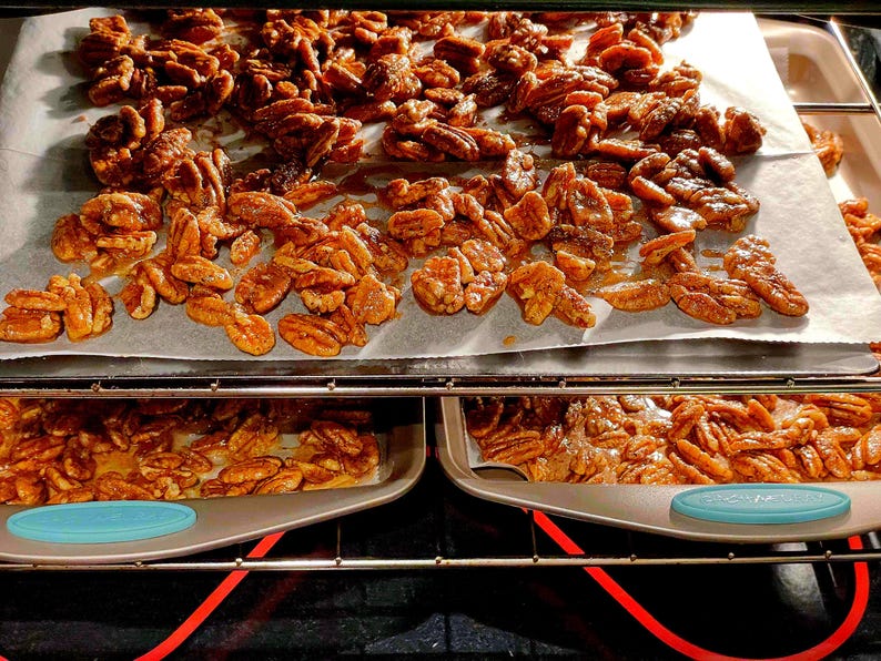 May include: Oven-baked pecans on parchment paper, with more pecans on trays below. The nuts are golden brown and appear glazed, suggesting a sweet coating. The oven's interior is visible, with red heating elements.