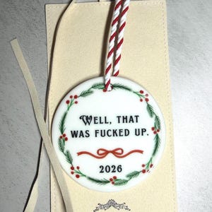 May include: A round white ceramic ornament featuring a green pine wreath with red berries and a red ribbon. The text reads "WELL, THAT WAS FUCKED UP." and "2026". It hangs from a red and white twisted cord and is placed on a light beige rectangular card.