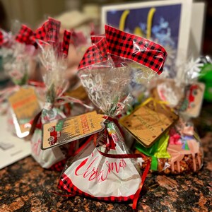 May include: Several cellophane wrapped gift bags with red and black buffalo plaid bows. The bags are tied with red ribbon and have tags attached. The bags have the word "Christmas" written on them in red script.