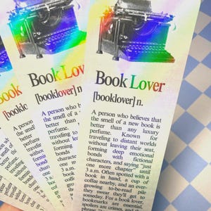 May include: Bookmarks with a vintage typewriter illustration and the words "Book Lover" in a colorful design. Each bookmark includes a definition of a book lover. The bookmarks are held in a hand.