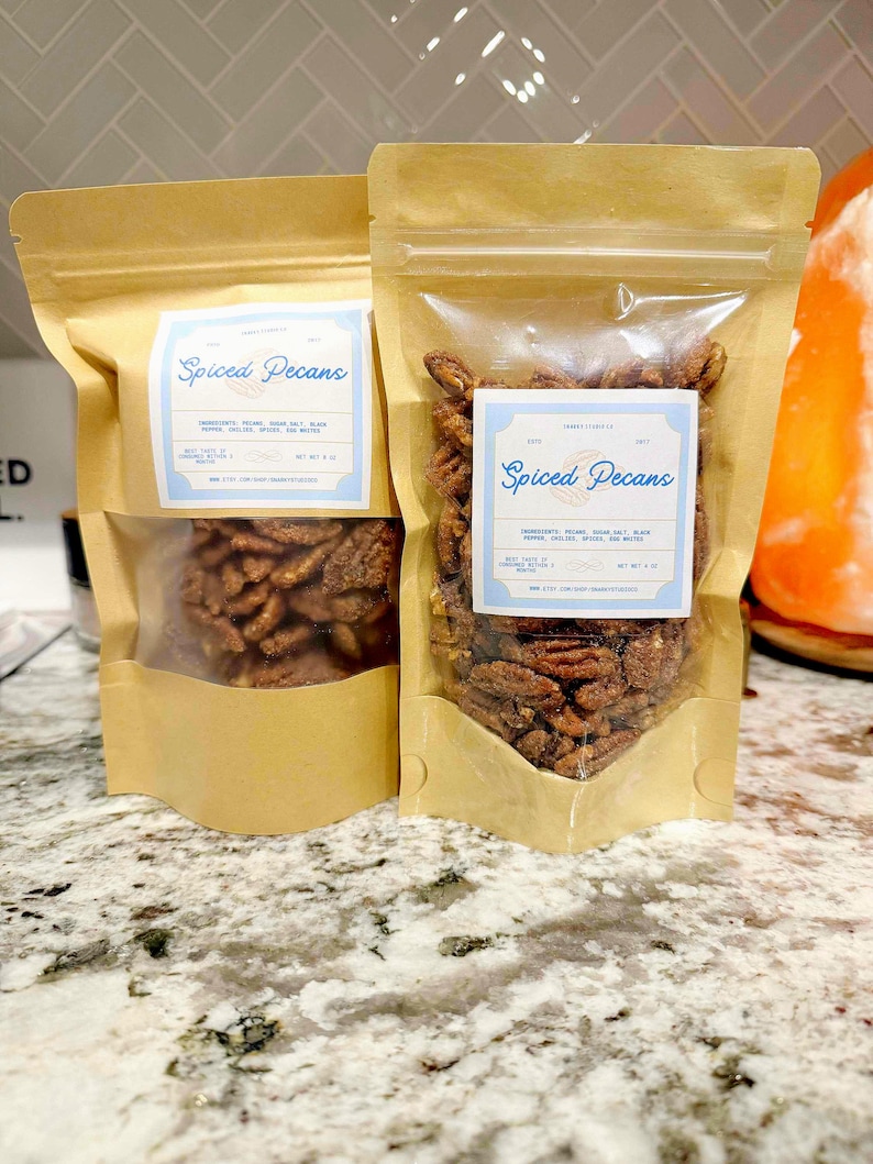 May include: Two kraft paper pouches filled with spiced pecans. Each pouch has a clear window showing the nuts. The pouches have a light blue label with the words "Spiced Pecans" in a script font. The ingredients are listed below.