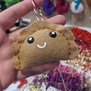 May include: A tan felt pierogi ornament with a smiling face, black button eyes, and orange stitching. It hangs from a red and white string. Other felt Christmas ornaments are visible in the background, including a snowman and dinosaurs.