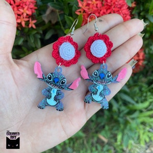 以下が含まれることがあります： 赤と白のクロシェの花のアクセントが付いた青いスティッチキャラクターのイヤリングのペア