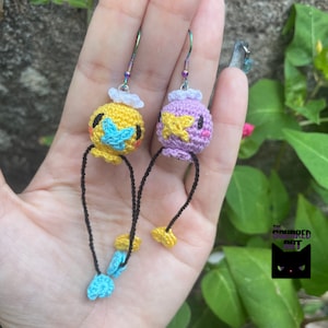 May include: A pair of handmade crochet earrings featuring whimsical, round characters. One earring is yellow with blue accents, the other is purple with yellow accents. Both have black dangling cords with blue and yellow details, and iridescent ear wires.