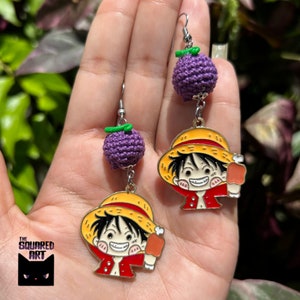 May include: Purple crocheted fruit earrings with a metal charm of a cartoon character wearing a straw hat, red shirt, and holding a meat bone.