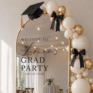 Peut inclure: Un miroir doré avec le texte "Welcome to Lauren's Grad Party Class of 2026". Le miroir est orné d'une coiffe de graduation noire et d'une arche de ballons blancs et dorés avec des nœuds noirs. Le miroir reflète une fête.