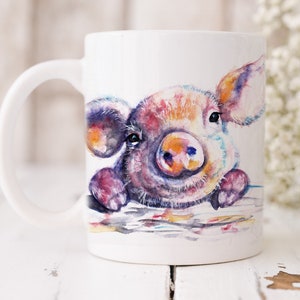 Könnte beinhalten: Weiße Keramiktasse mit einer Aquarellillustration eines bunten Schweins. Das Schwein hat eine rosa Schnauze, große Ohren und ist in Schattierungen von Lila, Orange und Blau gemalt. Die Tasse hat einen gebogenen Griff.