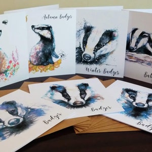 May include: A set of 10 watercolor greeting cards featuring various illustrations of badgers in different seasons. The cards are blank inside and perfect for sending a special message. The cards are arranged on a wooden table with brown envelopes.
