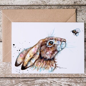 May include: Watercolor painting of a brown hare looking up at a bumblebee with black and yellow stripes. The hare has a white nose and brown fur with hints of blue and yellow. The background is white with black paint splatters.