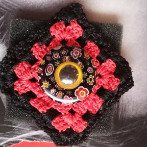 Peut inclure: Un carré crocheté noir et rouge avec un bouton coloré au centre. Le bouton a un fond noir avec des fleurs colorées et un cercle jaune au centre.