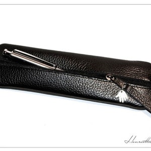 May include: A black leather pencil case with a zip closure and a silver charm. The case is empty, but a silver pen is partially sticking out of the zip opening.