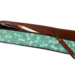 Op de afbeelding: Een bruine leren potlood etui met een ritssluiting en een stofvoering met een libellenpatroon. De etui heeft een zilveren hanger aan de rits.