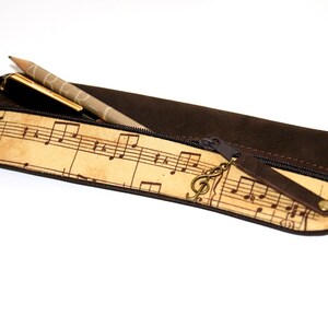 May include: A brown pencil case with a musical note pattern on the front. The case has a zipper closure and a small treble clef charm. A pencil and pen are partially visible inside.