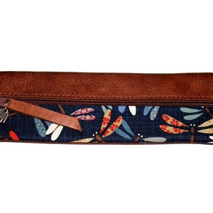 May include: A brown leather zippered pencil case with a blue fabric lining featuring a dragonfly pattern. The zipper pull has a small metal charm.