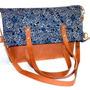 Könnte beinhalten: Eine braune Ledertasche mit einem blauen und weißen Blümchenmuster-Klappdeckel. Die Tasche hat zwei obere Henkel und einen langen Schultergurt.