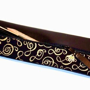 May include: A black pencil case with a gold musical note pattern. The case is partially unzipped, revealing a pencil and a pen. The zipper pull is brown. The top of the case is a solid dark brown color.