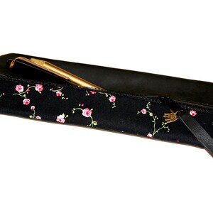 Op de afbeelding: Zwarte potlood etui met een ritssluiting en een klein roze bloemenpatroon. De etui heeft een zwarte leren band met een gouden metalen sluiting.