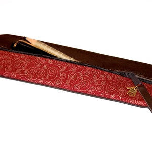 Può includere: Un astuccio per matite rosso con un motivo a spirale dorata e una zip in pelle marrone. L'astuccio è aperto e contiene una penna.