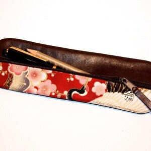 Op de afbeelding: Een bruine leren potlood etui met een ritssluiting. De etui is gevoerd met rode stof met een bloemenpatroon met witte kranen en gouden accenten. De rits heeft een metalen hanger met de tekst "Hand Made".