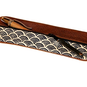 Könnte beinhalten: Eine braune Lederstiftetui mit Reißverschluss. Das Etui ist mit einem weißen Stoff mit schwarzem Wellenmuster gefüttert. Ein Stift befindet sich im Etui.