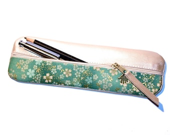 May include: A gold and green pencil case with a zipper closure and a small charm. The case is lined with a green fabric with a floral pattern and has two black pens inside.