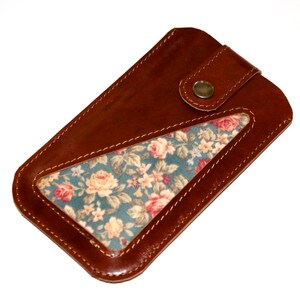 May include: A brown leather phone case with a floral fabric window. The case has a snap closure and stitched edges. The floral pattern features pink and yellow roses on a teal background.