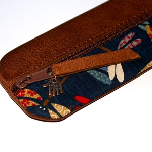 May include: A brown leather pencil case with a zipper closure and a blue fabric lining with a colourful floral pattern. The zipper pull has a metal charm that says "Hand Made".
