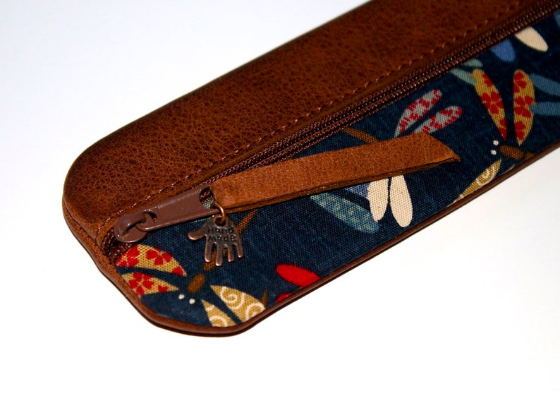 May include: A brown leather pencil case with a zipper closure and a blue fabric lining with a colourful floral pattern. The zipper pull has a metal charm that says "Hand Made".