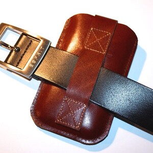 Könnte beinhalten: Ein braunes Leder-Handy-Etui mit einem schwarzen Ledergürtel. Der Gürtel hat eine silberne Schnalle mit der Gravur "Tribe".