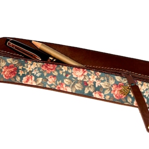 Könnte beinhalten: Ein braunes Leder-Federmäppchen mit Reißverschluss und einem floralen Stofffutter. Das Etui hat einen kleinen goldenen Anhänger am Reißverschluss.