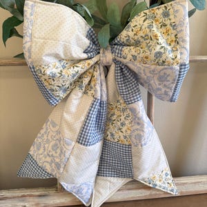 May include: A large, quilted bow in shades of white, blue, and yellow. The bow features a patchwork design with floral, gingham, and dotted patterns. It is likely used as a decorative element, possibly for a wreath or gift wrapping.