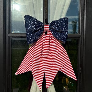 May include: A fabric bow decoration featuring a dark blue section with a white star pattern at the top and two long, pointed tails with a red and white horizontal striped pattern hanging below. The bow is secured with a central fabric band.