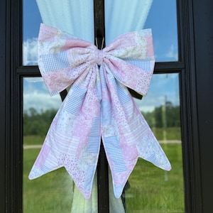 May include: A large, decorative bow made of patchwork fabric in shades of pink, white, and light blue. The bow is attached to a black window frame, with the outdoors visible through the glass.