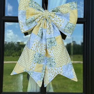 May include: A large, decorative bow made of patchwork fabric, featuring yellow gingham, floral, and blue patterned squares. The bow is tied in the center and hangs against a window with a black frame, reflecting the sky and greenery.