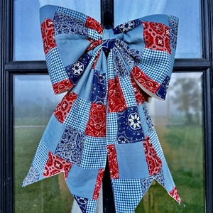May include: A large decorative bow constructed from a patchwork of fabric squares. The pattern features a mix of red, navy blue, and light blue bandana-style prints alongside small blue and white gingham checks, arranged in a repeating grid.