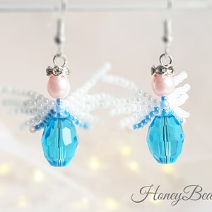 May include: A pair of beaded angel earrings with white wings, a light blue crystal body, and a pink pearl head. The earrings are hanging from silver hooks.