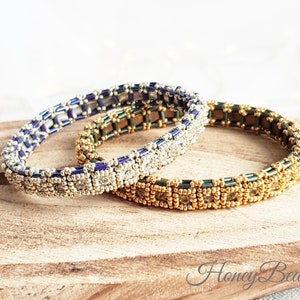 May include: Two beaded bracelets, one with silver and blue beads, the other with gold and green beads. The bracelets are stacked on top of each other on a wooden surface.