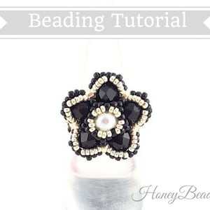 以下が含まれることがあります： 白い真珠の中心を持つ黒と金のビーズの花のリング。リングは白い背景の上にあります。 Honey Beads1.